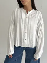 Blusa blanca con cuello mao y cierre frontal de botones de nácar. Presenta mangas largas abullonadas con puños elásticos y detalle de tablas verticales en el frente.