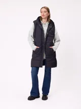 Chaleco largo acolchado color beige, con capucha forrada en sherpa, cierre frontal con cremallera y botones a presión, y bolsillos laterales.