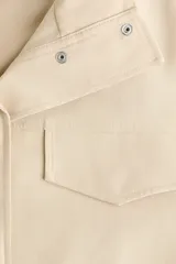 Campera beige con cuello chimenea, botones a presión ocultos en la parte delantera, bolsillos superiores con solapa y bolsillos laterales. Cuenta con diseño forrado y ajuste con cordón elástico en el bajo.