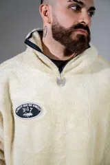 Canguro de peluche color crema, con cuello alto y cierre frontal parcial. Presenta un parche ovalado con diseño gráfico en el pecho y un bolsillo frontal tipo canguro.