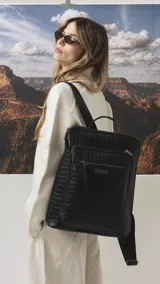 Mochila de diseño rectangular con textura símil cuero efecto cocodrilo en color tostado. Cuenta con un bolsillo frontal con cierre, compartimento principal con doble cierre, correas ajustables y manija superior. Incluye detalles metálicos y placa con logo de la marca.