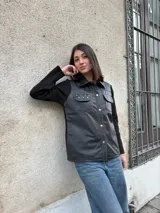 Campera negra con cuerpo de cuero y mangas de lana. Tiene corte recto, cuello camisero, cierre frontal con botones dorados, dos bolsillos con solapa en el pecho y detalles de tachas doradas.
