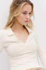 Top entallado corto de punto acanalado color blanco crema, con cuello camisero y escote en V. Tiene mangas largas.