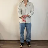 Camisa de manga larga color beige claro, con cuello clásico, cierre frontal con botones y logo bordado en el pecho. Corte regular.