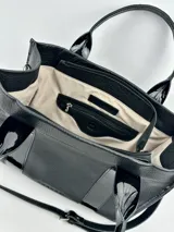 Bolso tote negro de cuero con detalles de charol, doble asa corta y correa bandolera de tela con letras.