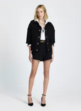 Chaqueta corta negra de tela tejida con cuello alto, mangas largas con puños abotonados, dos bolsillos delanteros con solapa y cierre frontal con botones.