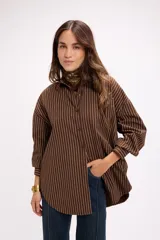 Camisa oversize de corte holgado con diseño de rayas verticales blancas sobre fondo marrón. Presenta cuello clásico, cierre frontal con botones y mangas largas con puños abotonados.