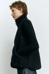 Chaleco de sherpa color negro con cuello elevado y cierre frontal. Presenta ribetes de cinta en los bordes, bolsillos tipo parche en la parte delantera y forro interior.