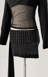 Conjunto de dos piezas compuesto por un blazer corto tipo torera de manga larga y una minifalda ajustada con detalle de strass brillante. La falda presenta un diseño drapeado y la chaqueta tiene un corte estructurado.