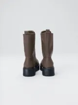 Botas de cuero color marrón de caña alta, con plataforma gruesa de goma y cierre metálico frontal con tirador. Presentan costuras a contratono y detalle de etiqueta de la marca en la lengüeta.