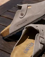 Zuecos de cuero gamuzado natural en color beige, con diseño de punta cerrada y talón descubierto. Presentan una plantilla anatómica de corcho y látex, con detalle de hebilla metálica grabada con el logo de la marca y nombre de la marca grabado en el lateral.