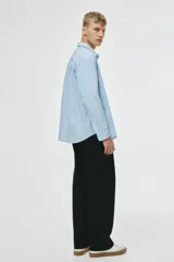 Camisa de popelina de algodón, color celeste pálido con finas rayas verticales negras. Presenta cuello inglés, tapeta clásica con botones blancos, bolsillo superior abierto en el pecho y canesú en la espalda. Mangas largas con puños abotonados y bajo levemente redondeado.