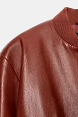 Chaqueta corta estilo bomber de Zara, confeccionada en material efecto piel color marrón rojizo. Presenta cuello redondo, hombreras, mangas abullonadas con puños elásticos y cierre frontal con cremallera oculta y botones a presión. Incluye bolsillos delanteros de vivo.