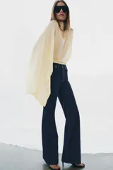 Jeans de tiro alto con corte flare y ajuste slim. Presenta trabillas en la cintura, bolsillos delanteros con detalle de botones metálicos y raya planchada a lo largo de las piernas. Cierre frontal con cremallera y botón.