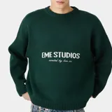 Suéter de punto grueso color verde oscuro, con corte relajado y ligeramente corto. Presenta el logo de la marca Eme Studios y la frase 'curated by ben ice' en el frente, realizados con técnica jacquard.