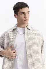 Camisa de popelina a cuadros, color beige y blanco, con cuello camisero, manga larga y bolsillo frontal. Presenta un estampado de letras en el pecho.