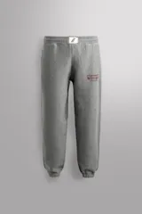 Pantalón jogger gris de corte regular fit, confeccionado en tejido de algodón con interior afelpado. Cuenta con cintura elástica ajustable con cordón, bolsillos laterales de vivo y traseros de plastrón. Presenta un texto estampado en color bordó en la pernera y bajo acabado en puño con elástico. Incluye un parche con logo combinado a contraste en la cintura.