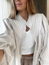 Camisa de algodón color beige con cuello mao, cierre frontal con botones y mangas abullonadas con frunces en los hombros. Presenta costuras verticales decorativas en el frente.