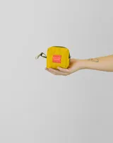 Monedero pequeño de forma cuadrada en color amarillo, con cierre superior y gancho metálico lateral. Presenta una etiqueta roja con el logo de la marca en el frente.