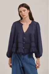 Camisa de color azul marino con cuello redondo y escote en V, mangas largas abullonadas con puños elásticos. Presenta un diseño con pliegues verticales en el pecho, detalles de encaje y volados en la parte inferior, con cierre frontal de botones.