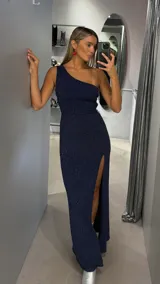 Vestido largo de fiesta color azul marino con acabado brillante y diseño de un solo hombro. Presenta un corte ajustado al cuerpo y una abertura lateral profunda en la falda.