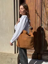 Cartera tipo tote bag de cuero color camel, con detalles de tachas metálicas en el bolsillo frontal y tiras largas para el hombro.