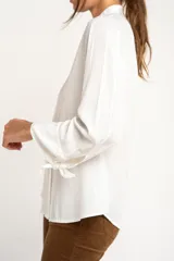 Camisa blanca de manga larga con cuello mao y cierre frontal con botones. Las mangas tienen nudos decorativos en los puños.