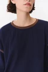 Remera de manga larga con corte oversized, color azul marino con cuello redondo en contraste color marrón. Presenta costuras decorativas a contratono hechas a mano en la zona de los hombros y logo bordado en el centro del pecho.