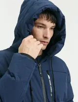 Campera puffer de corte largo, color verde militar, con capucha fija, cierre frontal y puños elásticos internos.