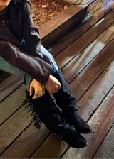 Bota alta de gamuza negra con caña alta, flecos laterales, tiras con hebillas metálicas en el tobillo y taco cuadrado.