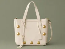 Bolso tipo tote color arena, confeccionado en cuero vacuno, con diseño estructurado y detalles de tachas metálicas doradas circulares. Cuenta con doble asa superior, correa larga desmontable y ajustable, cierre principal, bolsillo trasero con cierre y compartimentos internos.