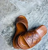 Sandalias tipo chinelas de cuero color suela, con plataforma baja y diseño de capellada con calados geométricos inspirados en motivos pampeanos.