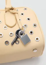 Bolso tipo bowling de color beige, adornado con múltiples ojales metálicos plateados en la parte frontal. Cuenta con doble asa de mano superior y cierre con cremallera.