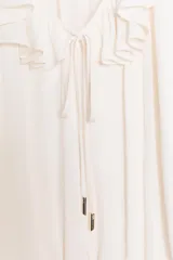 Blusa de georgette en color blanco, con escote en pico, detalle de volantes y lazada frontal. Presenta mangas largas abullonadas con puños ajustados y un bajo asimétrico con diseño tipo globo.