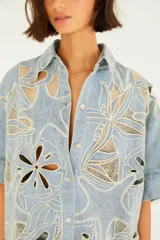 Camisa de jean de corte amplio con cuello camisero, mangas cortas y cierre frontal con botones. Presenta elaborados detalles calados y bordados.