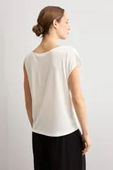 Blusa blanca de manga corta y corte recto, con escote bote y detalle metálico circular en el hombro.
