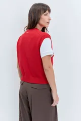 Chaleco de punto rojo con cierre frontal de botones, cuello redondo y dos bolsillos frontales con solapa. Presenta un diseño de tejido acanalado en los bordes y un pequeño parche decorativo en el pecho.