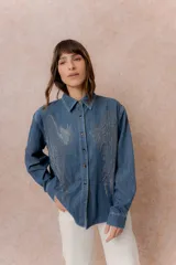 Camisa de denim liviano con calce relajado, cuello camisero clásico y cierre frontal con botones. Presenta una destacada aplicación de strass en el frente formando un diseño de alas.