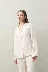 Camisa de pijama color blanco tiza con rayas verticales en relieve, de silueta clásica y caída fluida. Presenta cuello camisero, cierre frontal con botones y detalles de vivo rosado en cuello, puños y solapas.