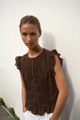 Blusa de cuello redondo y manga sisa con volantes, color marrón, con detalles de entredoses a tono, jaretas delanteras y cierre frontal con botones.