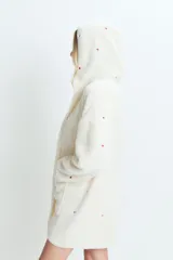 Saco de textura tipo peluche, color crema, con capucha y bolsillos frontales. Presenta un estampado sutil de corazones rojos dispersos por toda la prenda.