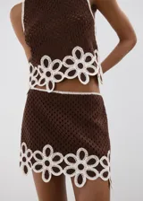 Falda corta de tejido crochet color marrón con detalles de flores blancas en el borde inferior. Silueta recta y forro interior.