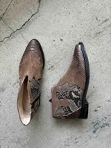 Bota corta de cuero color marrón, con taco cuadrado y punta fina. Presenta una correa con tachas pequeñas y una argolla metálica decorativa alrededor del tobillo. Cuenta con cierre lateral.