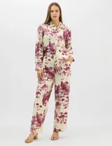 Conjunto de dos piezas compuesto por camisa de manga larga con cuello solapa y botones frontales, y pantalón de tiro alto con pierna recta y bolsillos laterales. Ambas prendas presentan un estampado paisajístico en tonos violeta sobre fondo crema.