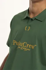 Remera de manga corta color verde oscuro, estilo polo con cuello y abertura frontal. Presenta un bordado central con el texto "PulauCrew" y un laurel en la parte superior.