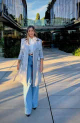 Trench coat gris de corte clásico con doble botonadura, solapas amplias, cinturón ajustable con hebilla y mangas con trabillas en los puños.