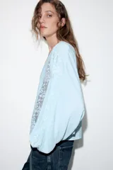 Blusa de escote en pico con cierre frontal de botones, mangas largas abullonadas con bordados a contraste y detalles de encaje tipo entredós y jaretas en el frente.