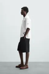 Camisa de manga corta confeccionada en mezcla de algodón y lino, color blanco. Presenta un cuello tipo bowling, calce regular y cierre frontal con botones. Destaca por sus bordados lineales en contraste de color negro con motivos de frutas y hojas distribuidos por toda la prenda.