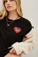 Remera negra de algodón con cuello redondo y manga corta. Presenta un aplique bordado en el pecho con forma de corazón rojo que contiene un ojo detallado con mostacillas y bordados.