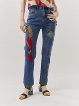 Pantalón de jean tiro alto tipo mom-fit, con parches de colores en forma geométrica, detalles de tientos de cuero trenzados en la cintura y adornos de canutillos en la parte frontal.
